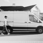 A delivery man walking past a panel van on a sunny residential street in monochrome.